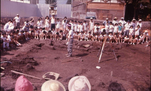 中央で調査員が説明を行っており、その周りで小学生が話を聞いています。